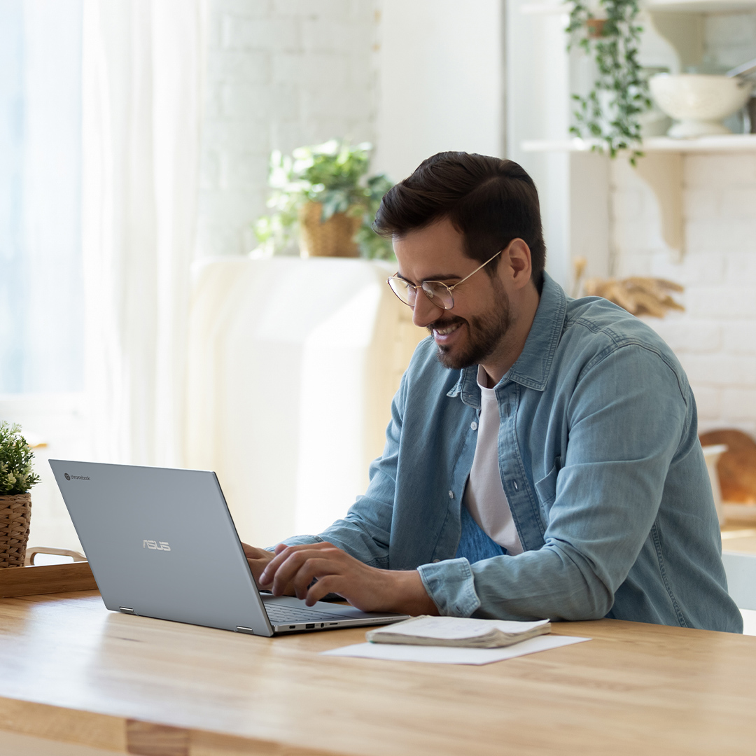 En vy av en man som ler medan han skriver på en ASUS Chromebook som står på vänster sida på ett träbord.